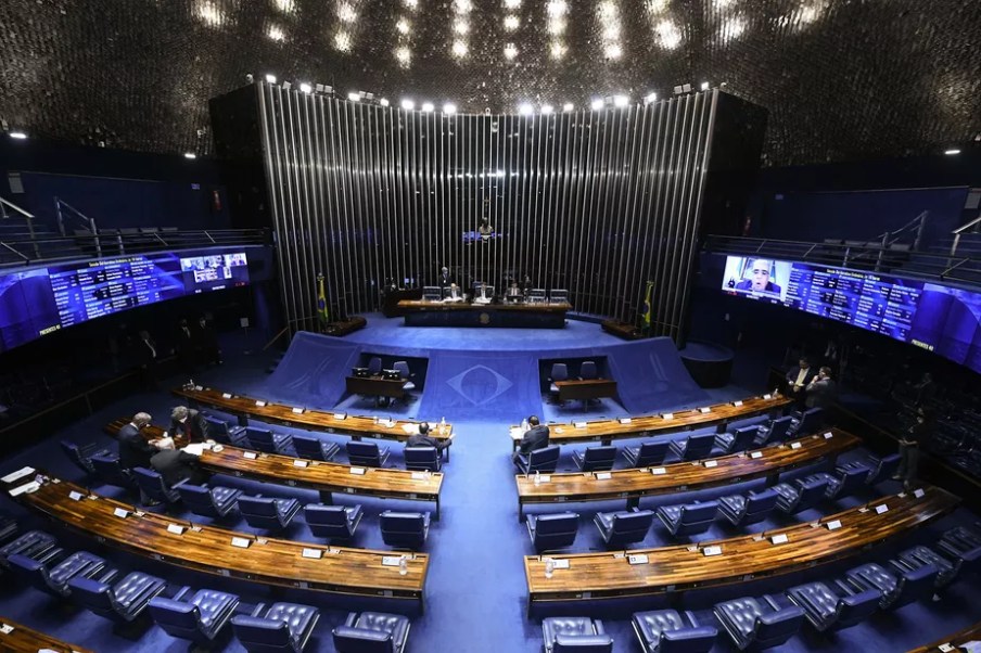 Foto do plenário do Senado Federal, com fileiras de cadeiras e microfones aos lados e a mesa diretora no centro