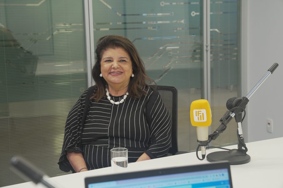 Foto de Luiza Trajano, mulher branca vestindo uma roupa preta com traços branco e um colar branco em um ambiente de escritório. Ela está sorrindo e à frente dela está um microfone amarelo com o símbolo da IF.