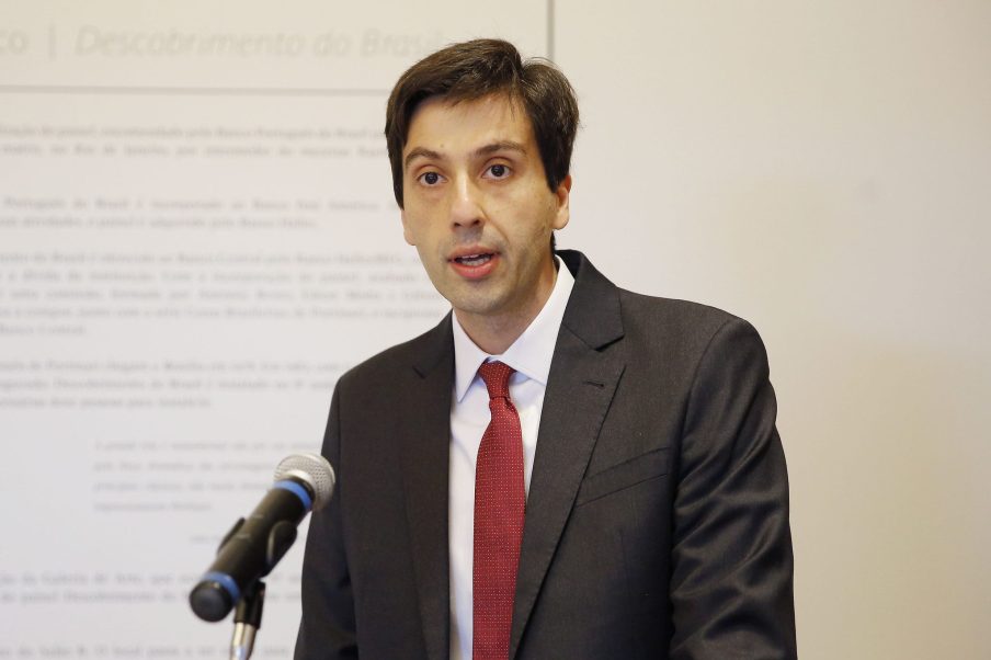 Foto de um homem branco usando um paletó escuro com uma gravata vermelha e camisa branca.