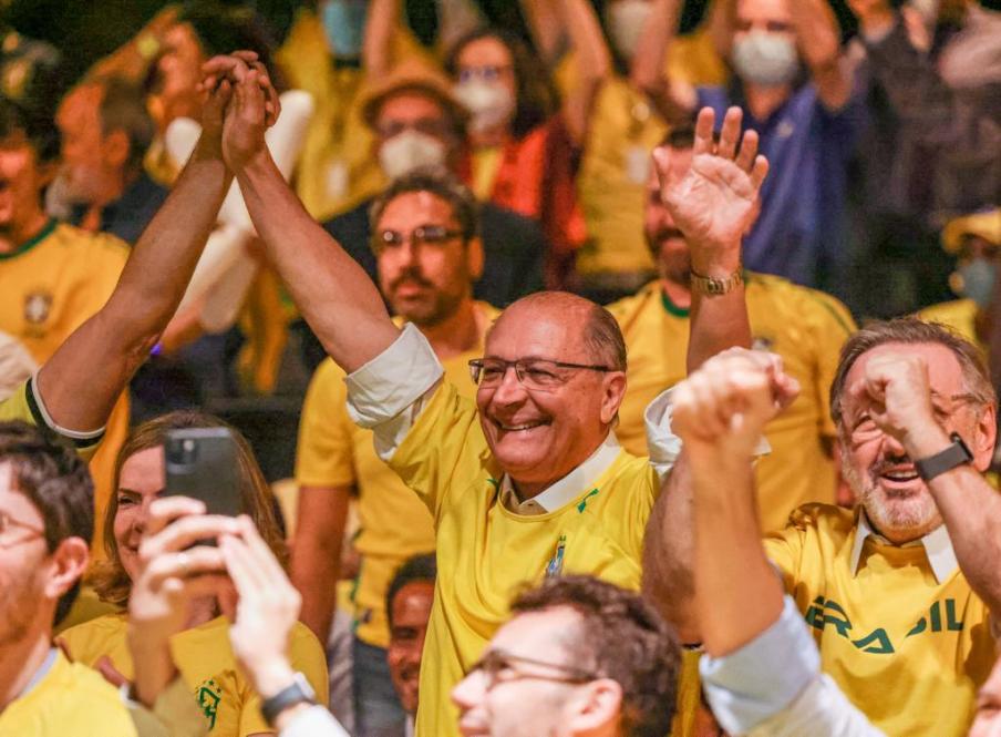 Foto de Geraldo Alckmin e de Gleisi Hoffman vestindo a camisa da seleção brasileira e comemorando um gol