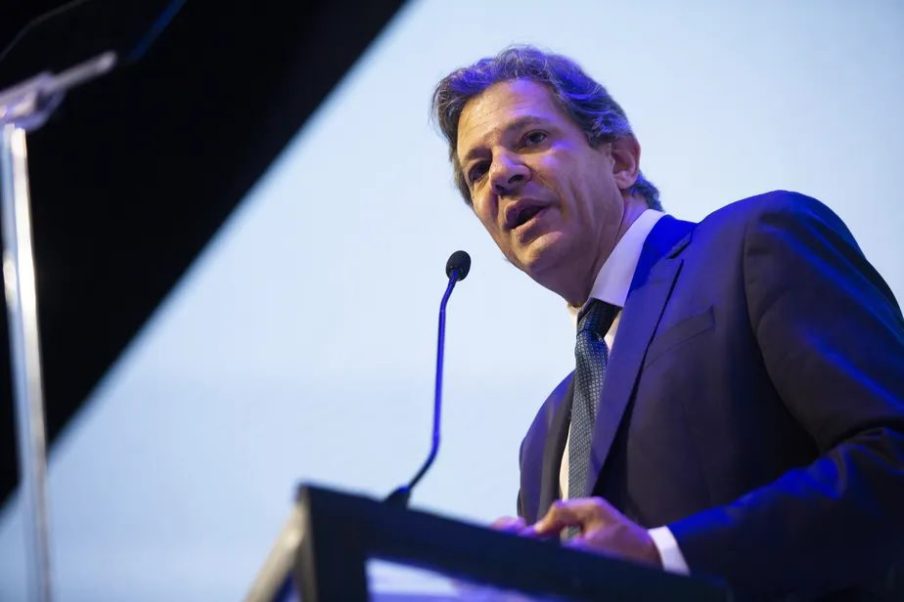 Foto de Fernando Haddad, ministro da Fazenda. Ele está atrás de um púlpito, usando gravata verde-escura e um terno cinza