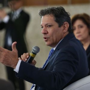 Foto de Fernando Haddad, ministro da Fazenda. Ele é homem, usa terno preto, camisa branca e gravata xadrez com listras brancas. Ele fala e gesticula com um microfone na mão direita