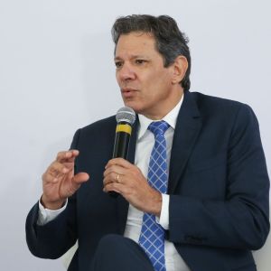 Foto de Fernando Haddad, ministro da Fazenda. Ele é homem, usa terno preto, camisa branca e gravata xadrez com listras brancas. Ele fala e gesticula com um microfone na mão esquerda