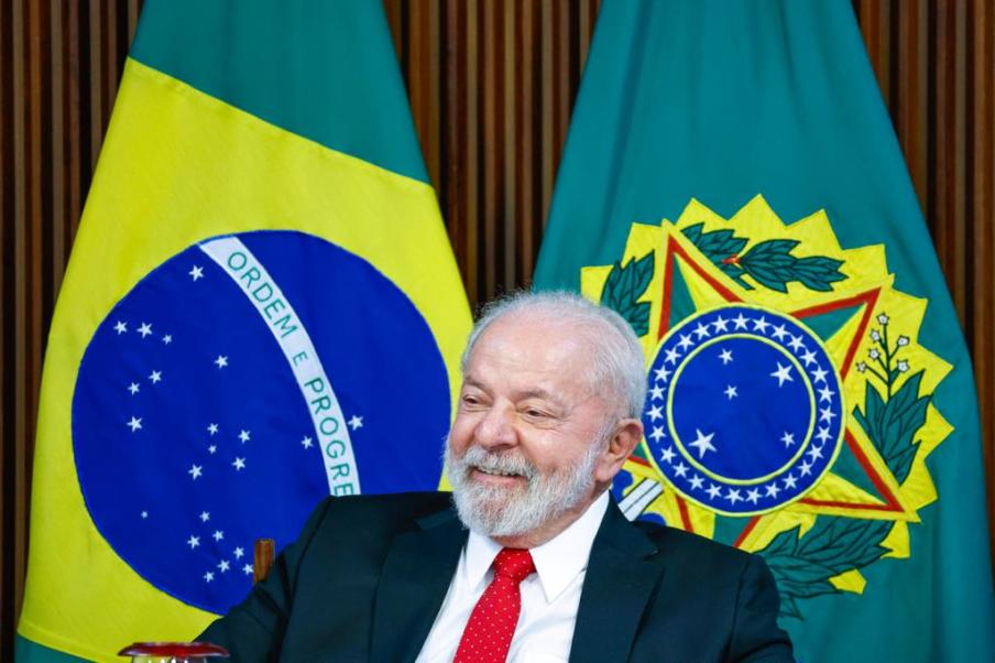 Foto de Luiz Inácio Lula da Silva, presidente da República, sorrindo. Ele veste um terno preto, camisa branca e grava vemelha. Ao fundo, uma bandeira do Brasil e uma bandeira com o brasão da Presidência da República