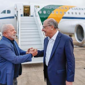 Foto de Luiz Inácio Lula da Silva, presidente do Brasil, cumprimentando Geraldo Alckmin, vice-presidente do Brasil. Eles se cumprimentam perante o avião presidencial com faixas verdes e amarelas. A escada de embarque está posicionada