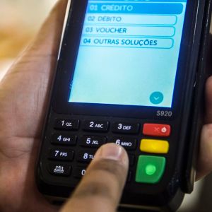 Foto de uma maquininha de cartão de pagamento coma opção de crédito selecionada. A mão do lojista está sobre a máquina