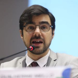 Foto de Guilherme Mello, secretário de Política Econômica da Fazenda. Ele é homem, tem cabelo castanho escuro e usa óculos.