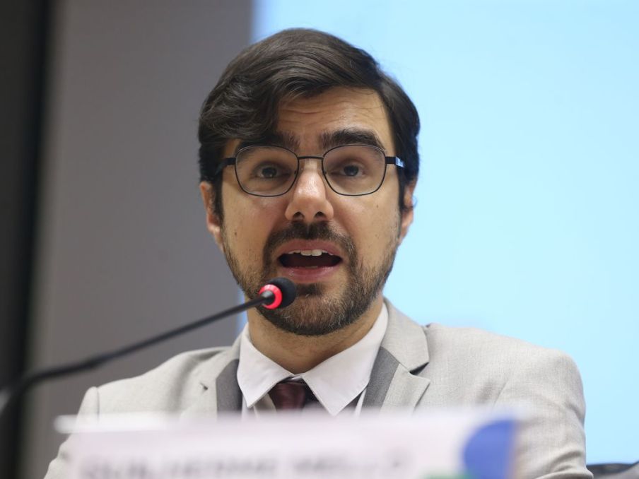 Foto de Guilherme Mello, secretário de Política Econômica da Fazenda. Ele é homem, tem cabelo castanho escuro e usa óculos.