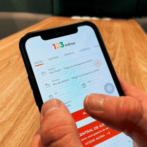 A imagem mostra uma mão segurando um celular, com o site da 123 milhas aberto na tela.