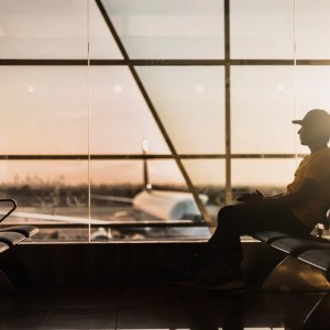 A imagem mostra um homem com boné sentado em uma cadeira dentro de um aeroporto. Ao fundo está um avião estacionado.
