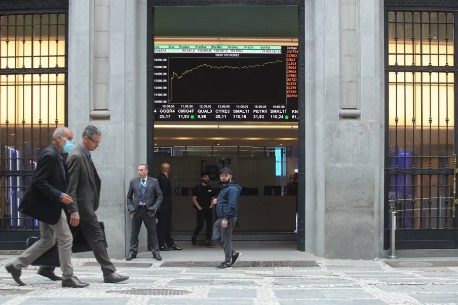 A imagem mostra a fachada da B3, bolsa de valores brasileira, com algumas pessoas passando na frente.