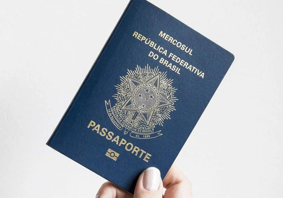 A imagem mostra uma mão segurando um passaporte brasileiro em um fundo branco.