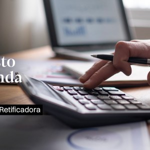Foto mostra apenas objetos e uma mão. Foco está em uma calculadora sendo operada pela mão de uma pessoa e ao fundo um computador. Os objetos estão sob uma mesa de madeira. Tema da matéria é Declaração de Imposto de Renda 2024