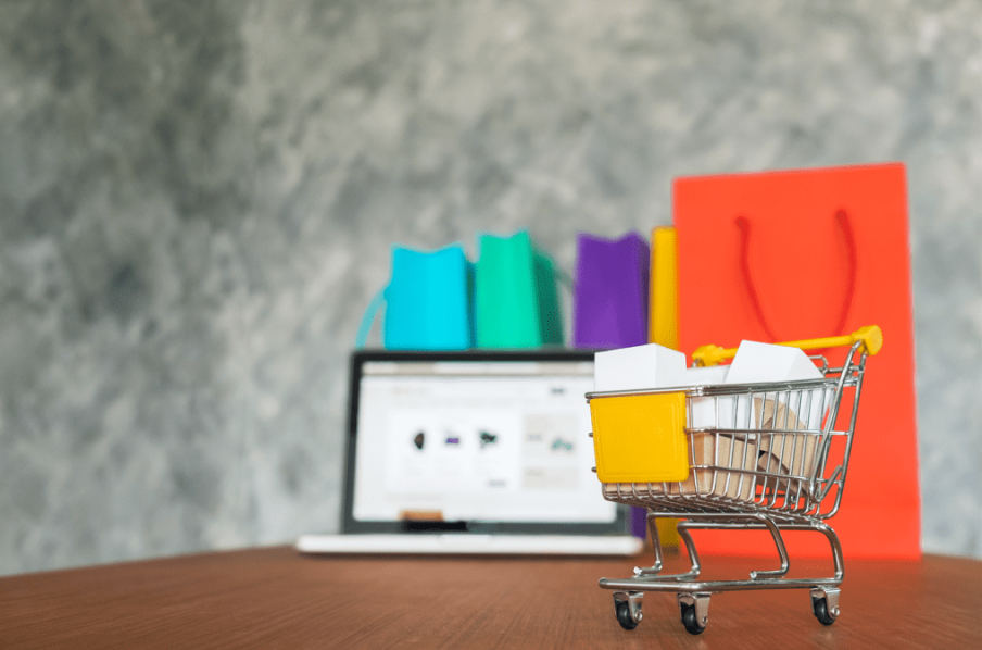 A imagem mostra um carrinho de compras com algumas sacolas dentro. Ao fundo um computador com outras sacolas de compras.