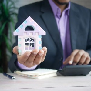 Um homem de paletó segurando uma casa em formato de maquete em uma mão e na outra fazendo contas em uma calculadora.
