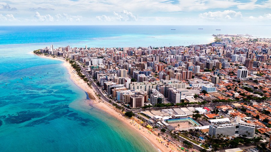 Foto de uma paisagem vista de cima de Maceió, em Alagoas. De um lado há um mar bem azulado. Do outro, uma cidade com prédios e casas.