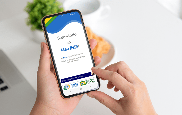 Imagem de uma mão segurando um celular. Na tela, está escrito "Bem-vindo ao Meu INSS".