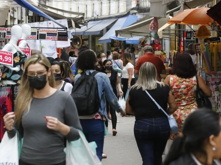 Foto de rua movimentada por pessoas com lojas ao redor. A matéria fala sobre investimentos em ações do setor de varejo na bolsa de valores.