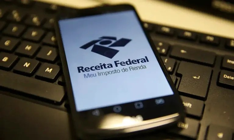 Foto mostra um celular sobre um teclado de computador. No visor do celular há o logotipo da Receita Federal do Brasil. Matéria fala sobre o tema: A partir de que idade se declara imposto de renda?