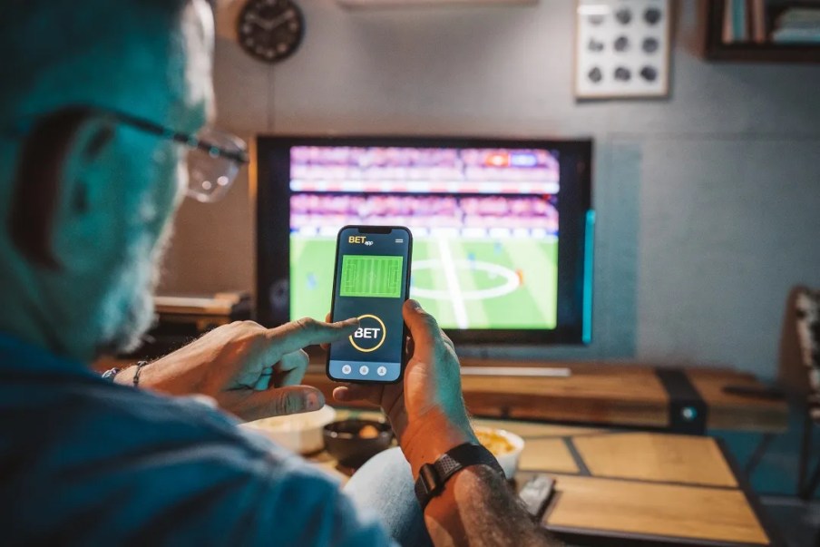 Foto de um homem branco segurando um celular, como se estivesse fazendo uma aposta esportiva. Ao fundo, uma televisão ligada em um jogo de futebol.