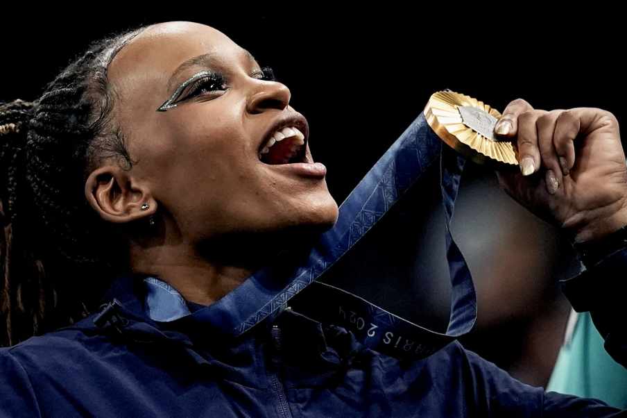 Foto da ginasta Rebeca Andrade levantando a medalha de ouro que ganhou nos Jogos Olímpicos de Paris, em 2024. Ela é mulher, negra.