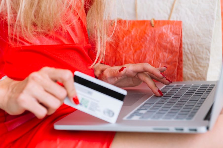 Imagem de uma mulher branca e loira fazendo uma compra pela internet segurando um cartão de crédito.