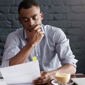 Imagem de um homem negro pensando e olhando para um papel. Ao lado, uma xícara de café.