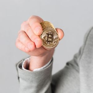 Foto de uma mão masculina segurando algumas moedas. No centro, há desenhado o símbolo do Bitcoin, com uma letra B.