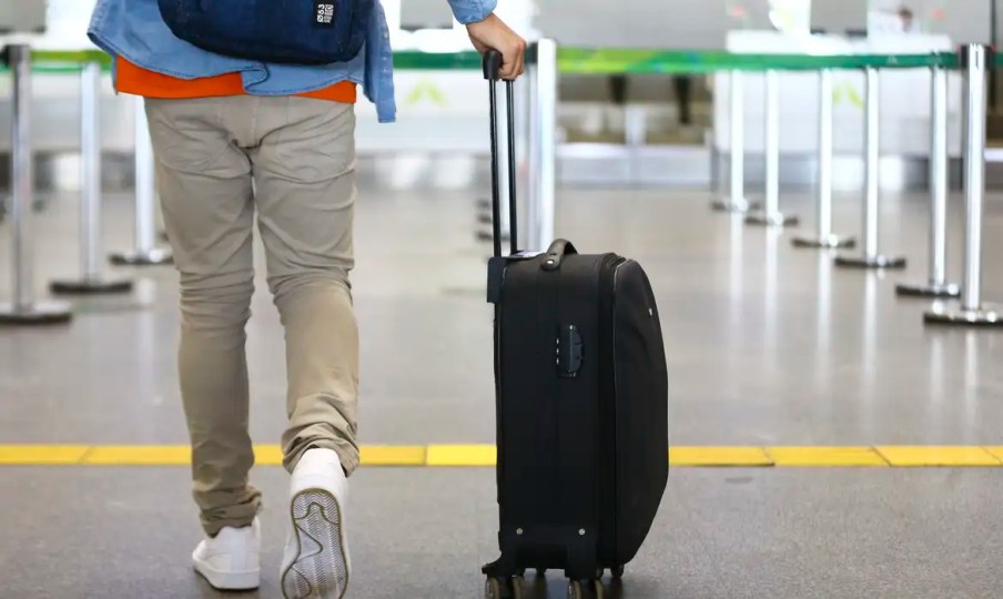 Imagem mostra passageiro, homem, branco, fotografado de costas, caminhando em aeroporto. Usa calça bege claro, camiseta vermelha e uma camisa jeans por cima. Arrasta com a mão direita uma mala de cor preta.