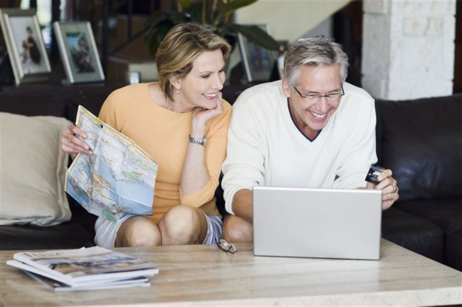 Foto de um casal mais velho olhando para um computador. Ela segura um mapa e ele um cartão de crédito.