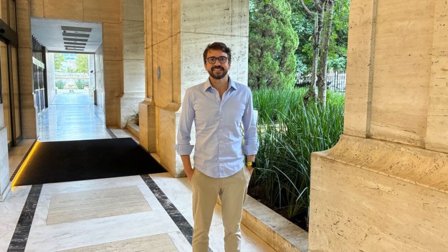 Imagem de um homem branco de camisa azul clara e calça bege com as mãos no bolso da calça sorrindo para a foto. Ele está em um prédio comercial.