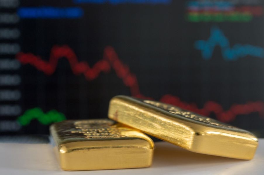 Imagem de duas barras de ouro e ao fundo gráficos do mercado financeiro.
