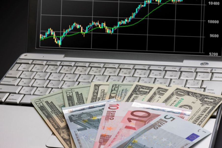 Imagem de um computador com gráfico na tela. Em cima do teclado, notas de dólar e euro.