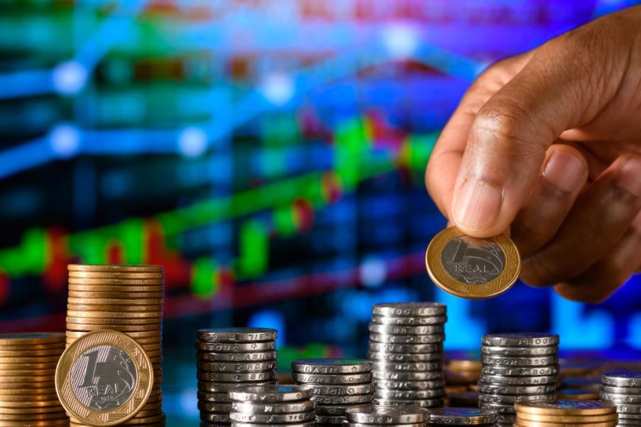 Imagem de uma mão empilhando moedas de 1 real e 50 centavos.
