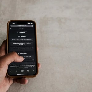 Imagem da mão de uma pessoa branca segurando um celular. Na tela está a plataforma de inteligência artificial ChatGPT.