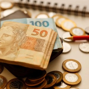 Imagem de um bolo de notas de 50 reais por cima de um bolo de notas de 100 reais. Ambos estão por cima de uma carteira preta. Ao redor, várias moedas de 1 real e 50 centavos. Junto a esses objetos tem um lápis vermelho e um caderno.