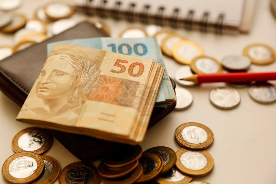 Imagem de um bolo de notas de 50 reais por cima de um bolo de notas de 100 reais. Ambos estão por cima de uma carteira preta. Ao redor, várias moedas de 1 real e 50 centavos. Junto a esses objetos tem um lápis vermelho e um caderno.